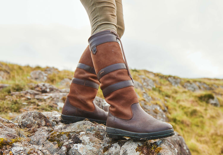 A side angle of the Dubarry Ladies knee-high walnut brown leather Galway Country Boot with laced top worn by model on a boulder in a grassland area.