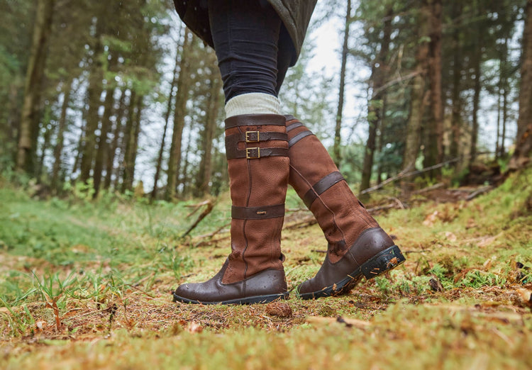 The side angle of Dubarry knee-high walnut brown leather Longford country Boot with side buckle detail in a grassland area.