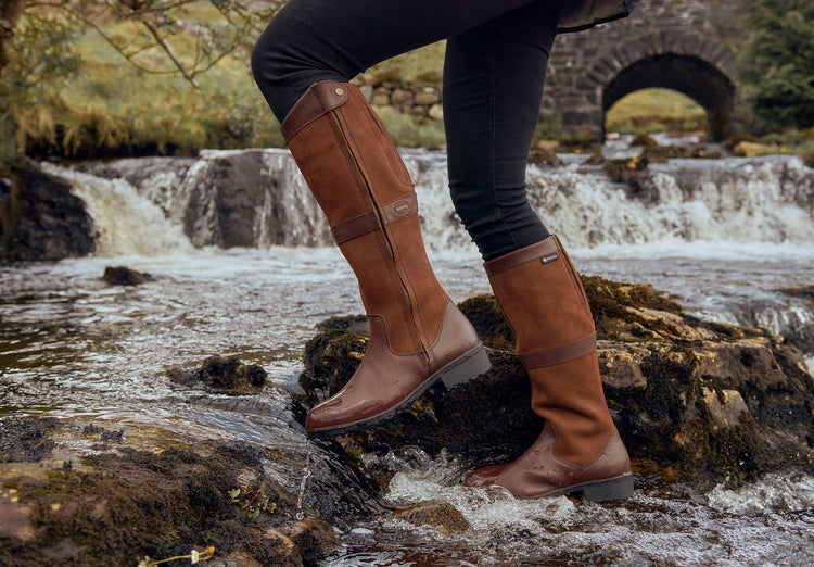 An on Model image of a pair of Dubarry walnut brown leather knee-high Sligo country boots with full zip walking through a river bank.