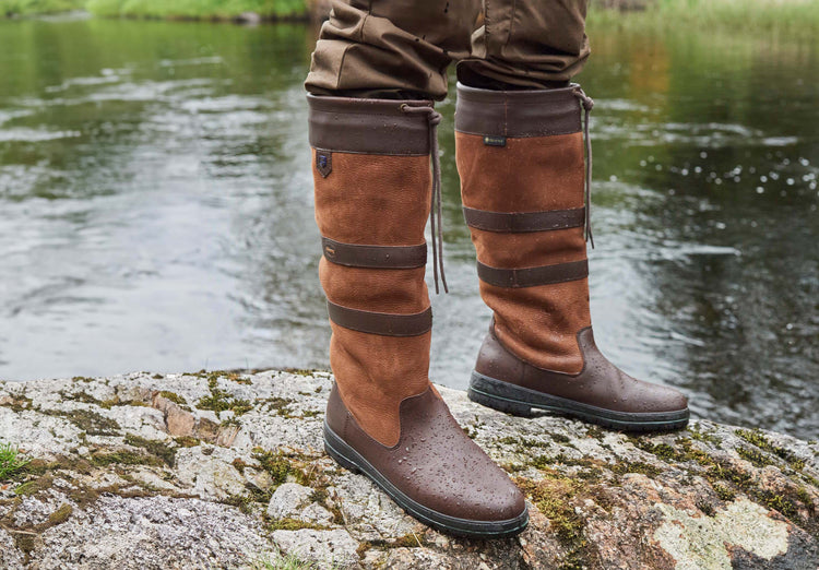 Side angle of the Dubarry Men's knee-high walnut brown leather Galway Country Boot with laced top worn by model on a boulder next to a lake.