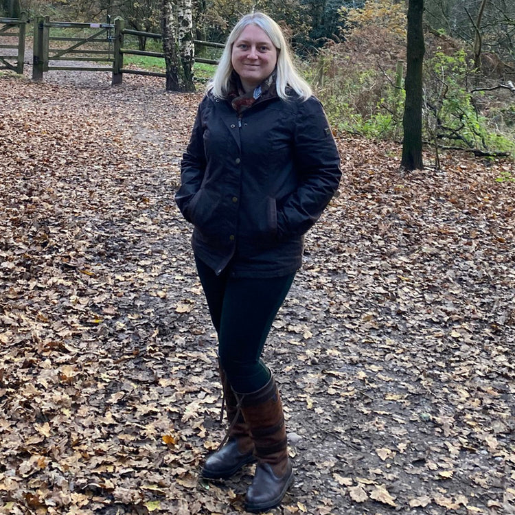 A square image of a woman wearing a Mountrath Wax Jacket in a forest in an autumnal setting with brown leaves littering the floor.//© Outdoor & Country